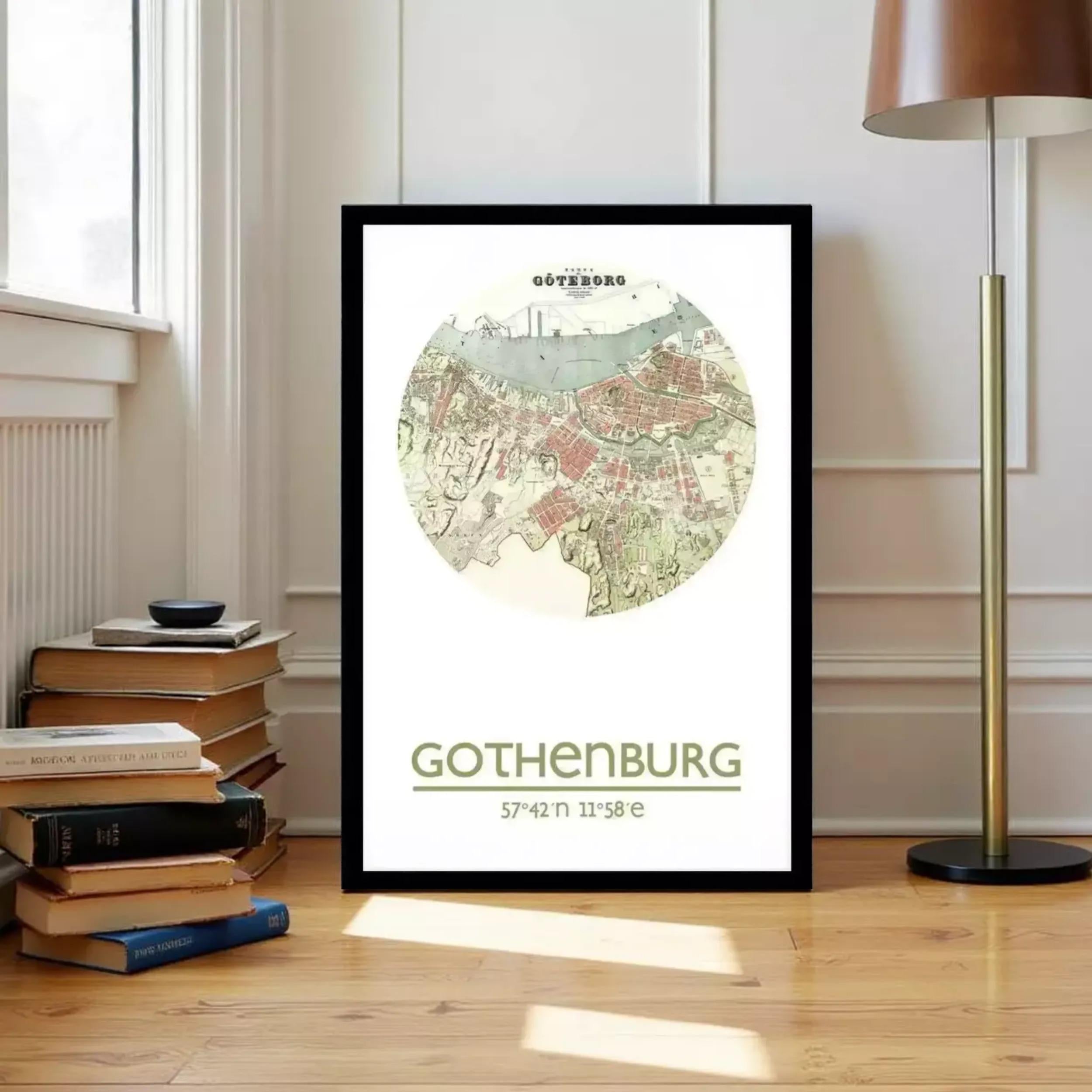 A framed map of Gothenburg, a city in Sweden, is displayed on a wooden floor next to a stack of books and a lamp.