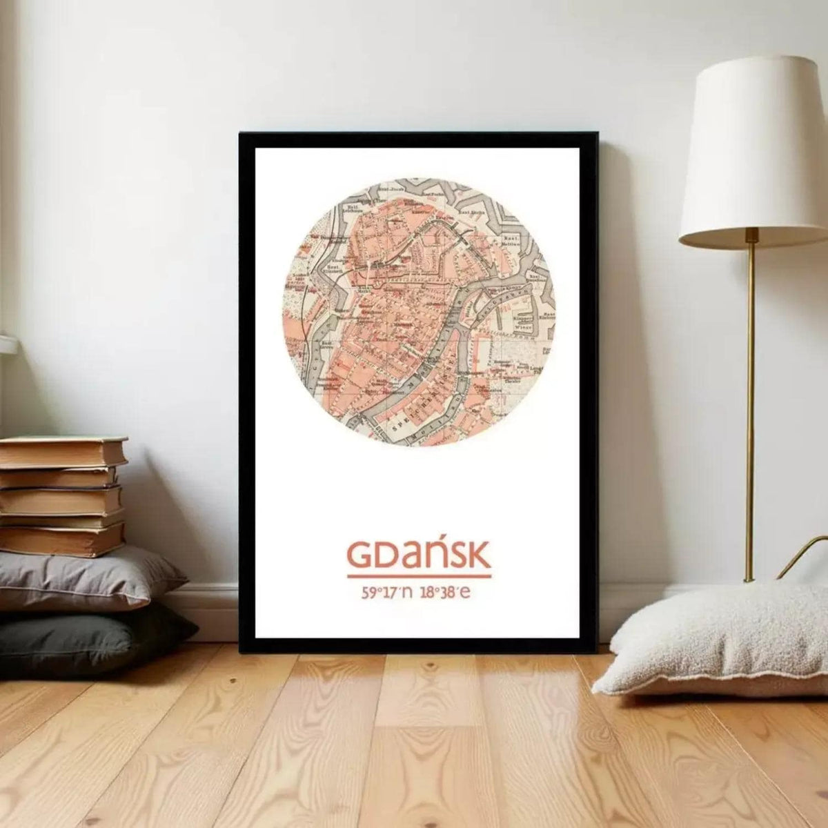 A framed map of Gdansk, Poland, with the city's name and location displayed prominently. The map is surrounded by a circular design, and the text "Gdansk" is written below it. The image is set against a white wall, with a lamp and some books visible in the background.