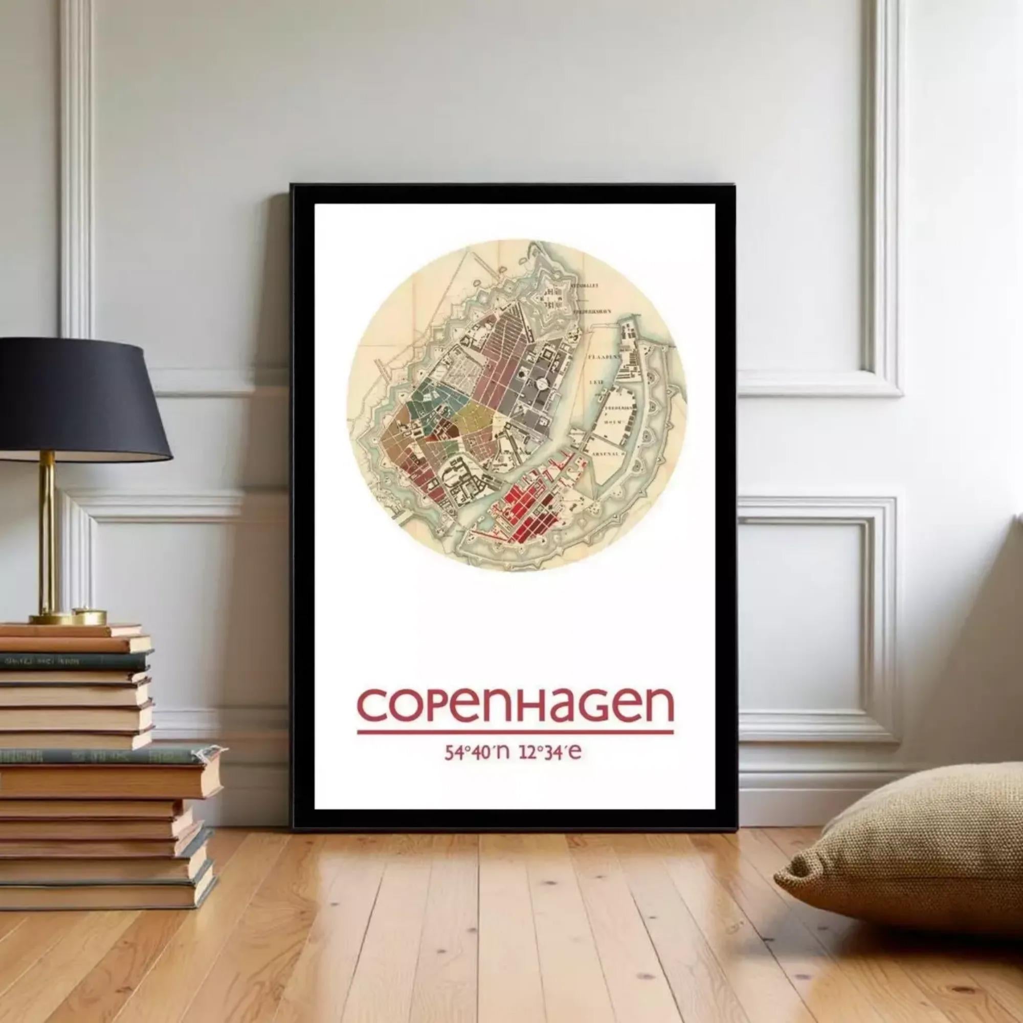A framed poster of a map of Copenhagen, Denmark, with the text "COPENHAGEN" and "54°N 12°34°E" printed on it, displayed on a wall in a room with a stack of books and a lamp.