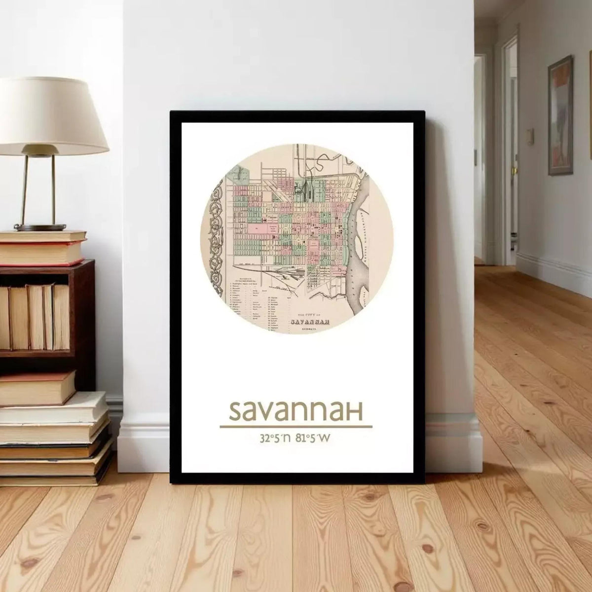 A framed map of Savannah, Georgia, with the text "Savannah Georgia" and "32.25°N 81.5°W" printed on it, standing on a wooden floor next to a lamp and books.