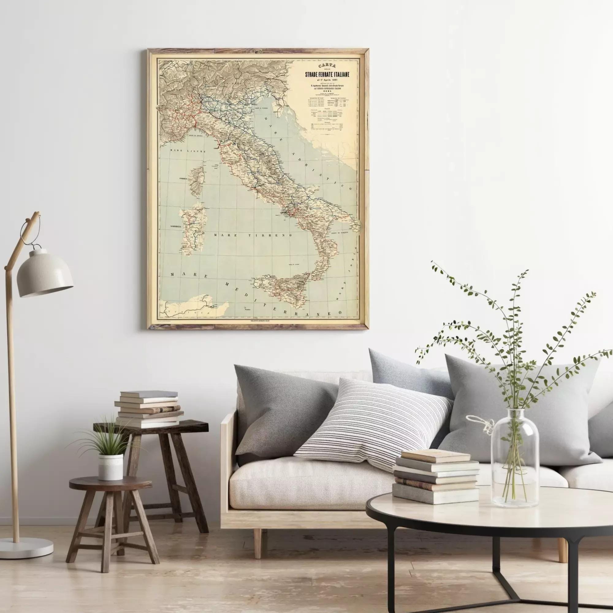 a living room with a large framed map of Italy hanging on the wall above a couch with pillows. There are also some books and a vase with greenery on the couch, creating a cozy and inviting atmosphere.