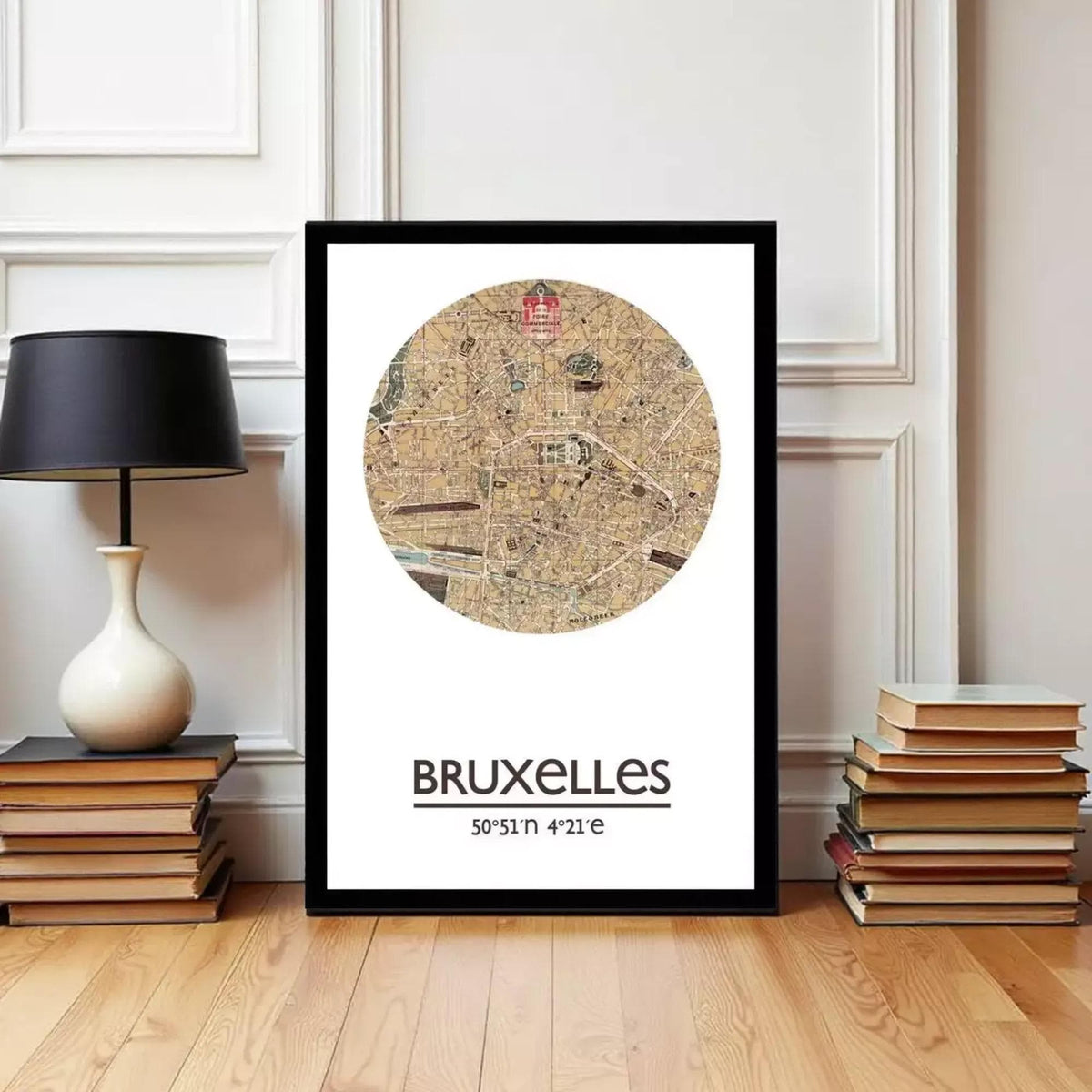 A framed map of Bruxelles, Belgium, with a black border and a circular design depicting the city's streets and landmarks. The map is placed on a wooden floor, with a stack of books and a black lamp to the left and a stack of books to the right.