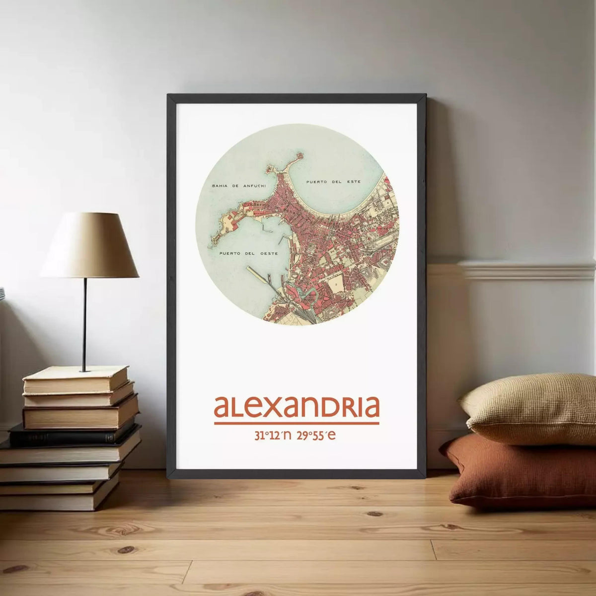 A framed map of the city of Alexandria, with the name "Alexandria" printed below it. The map is displayed on a wall, and there are some books and pillows visible in the foreground.
