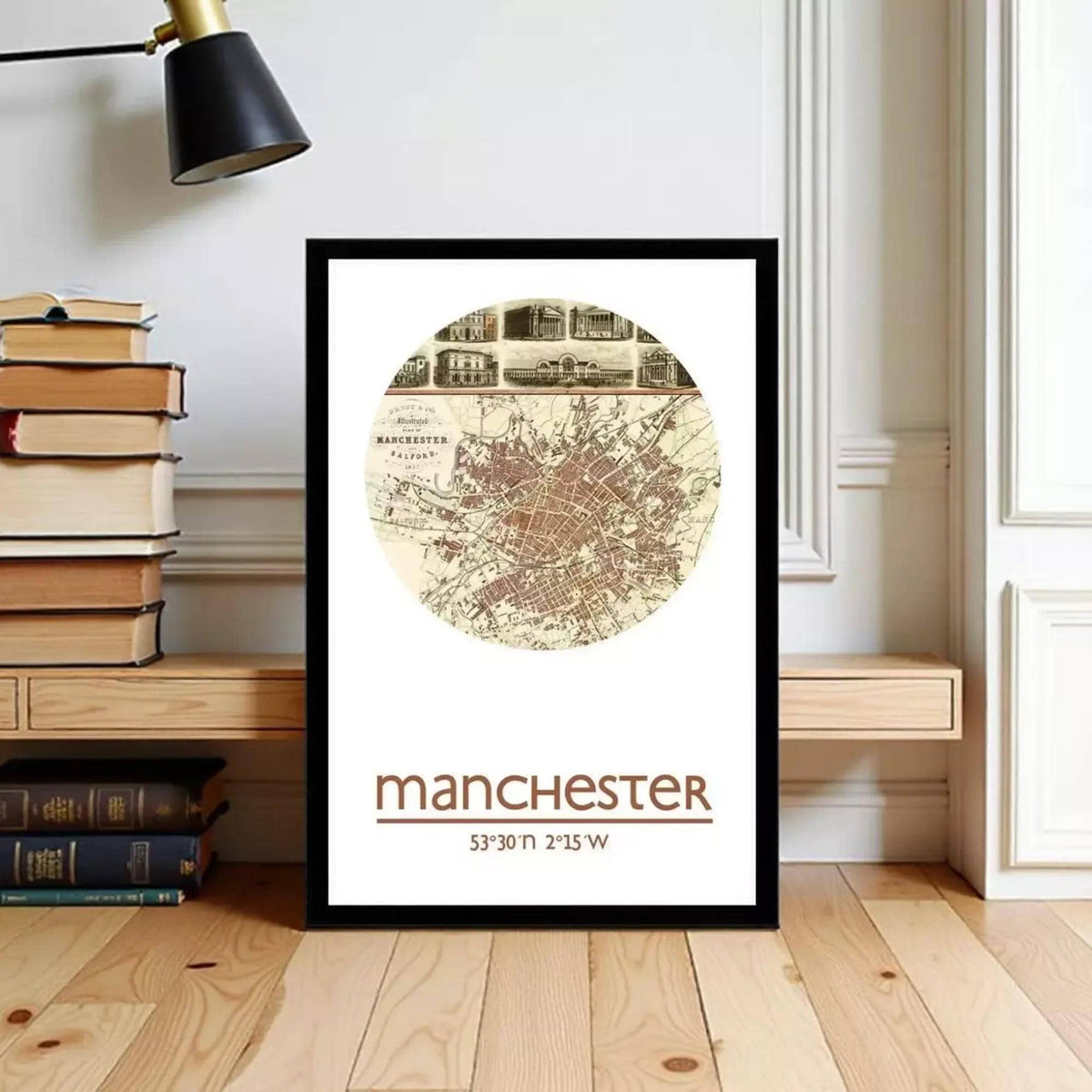 A framed map of Manchester, UK, with the text "Manchester" and "53°30'N 2°15'W" printed on it. The map is displayed on a wooden floor, with a stack of books and a lamp visible in the background.