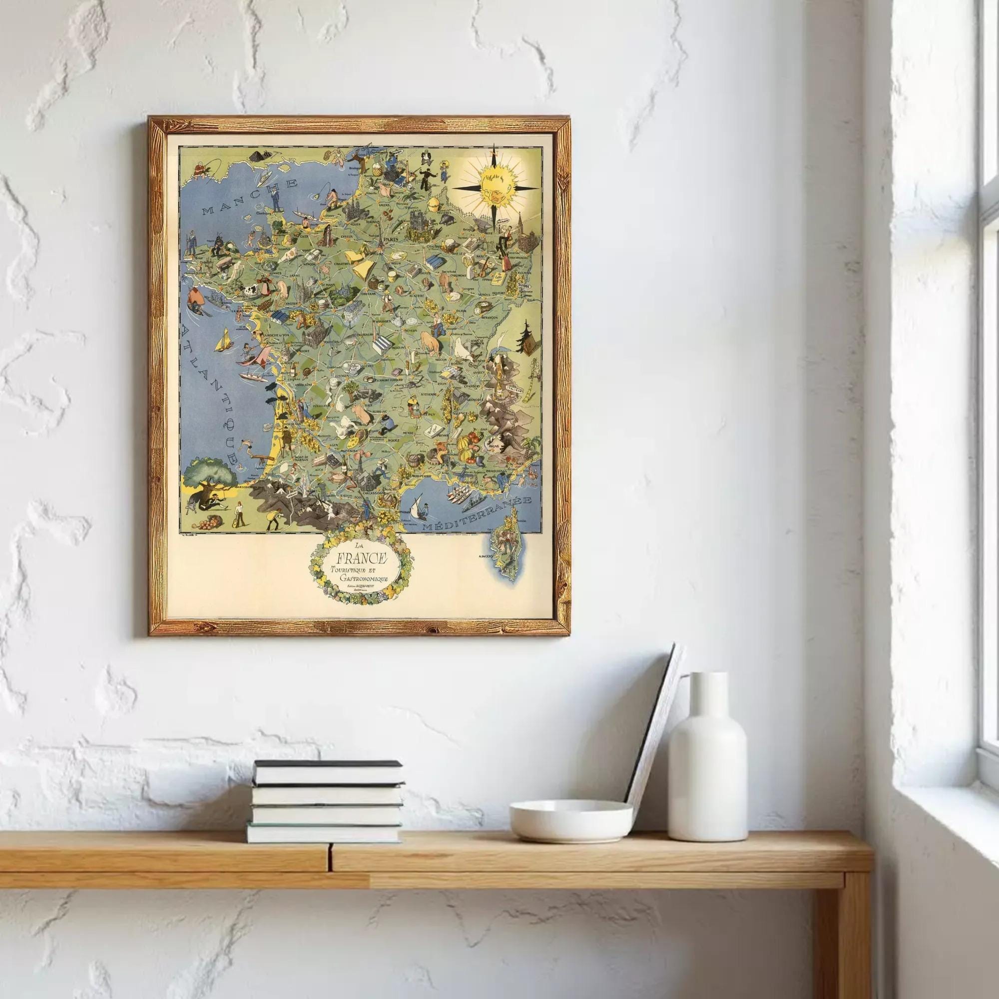 a framed map of France hanging on a wall, with a wooden shelf below it holding a stack of books and a white vase.