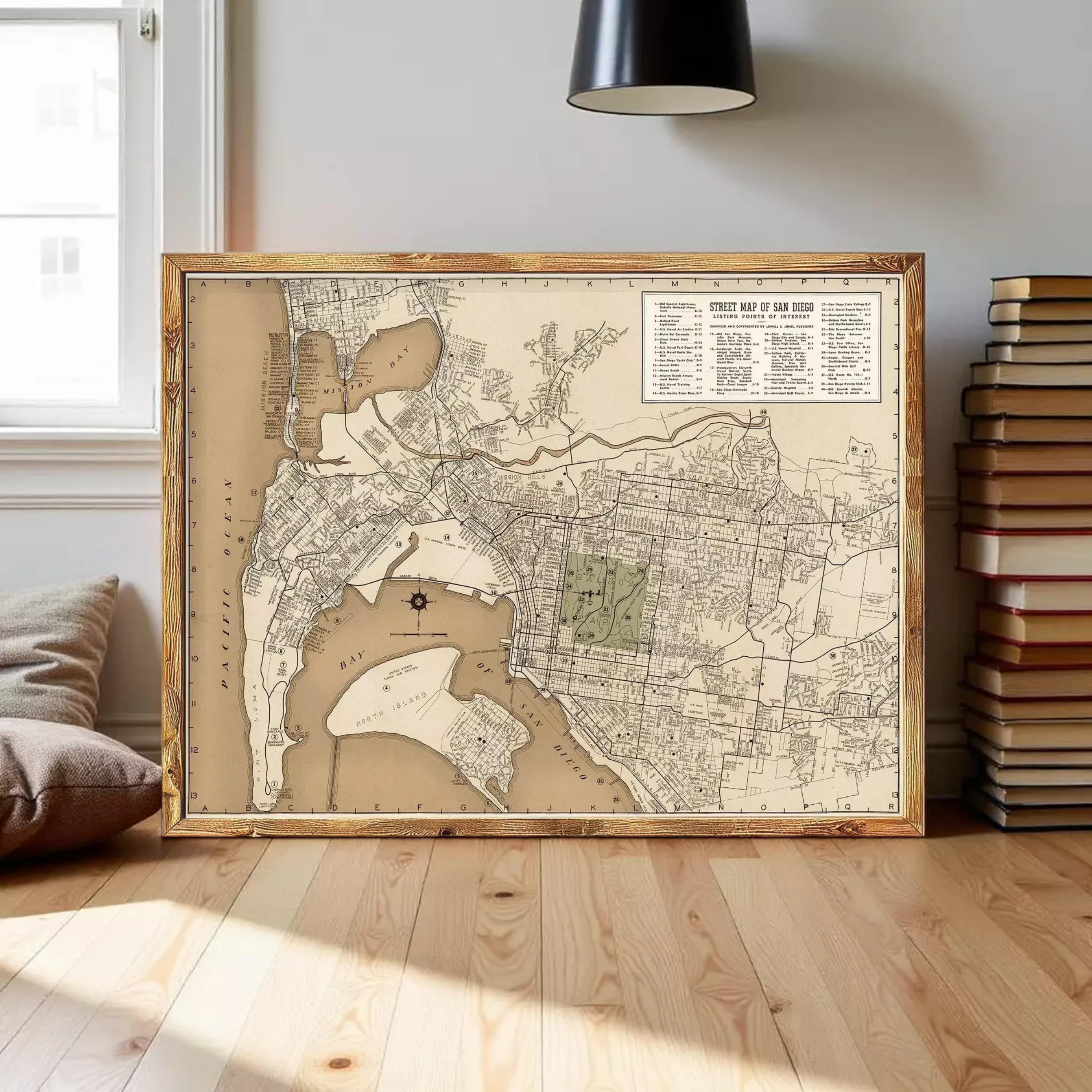 a large framed map of San Diego, California, with a wooden frame and a vintage style. The map is displayed on a wooden floor in a room with a window and a stack of books.