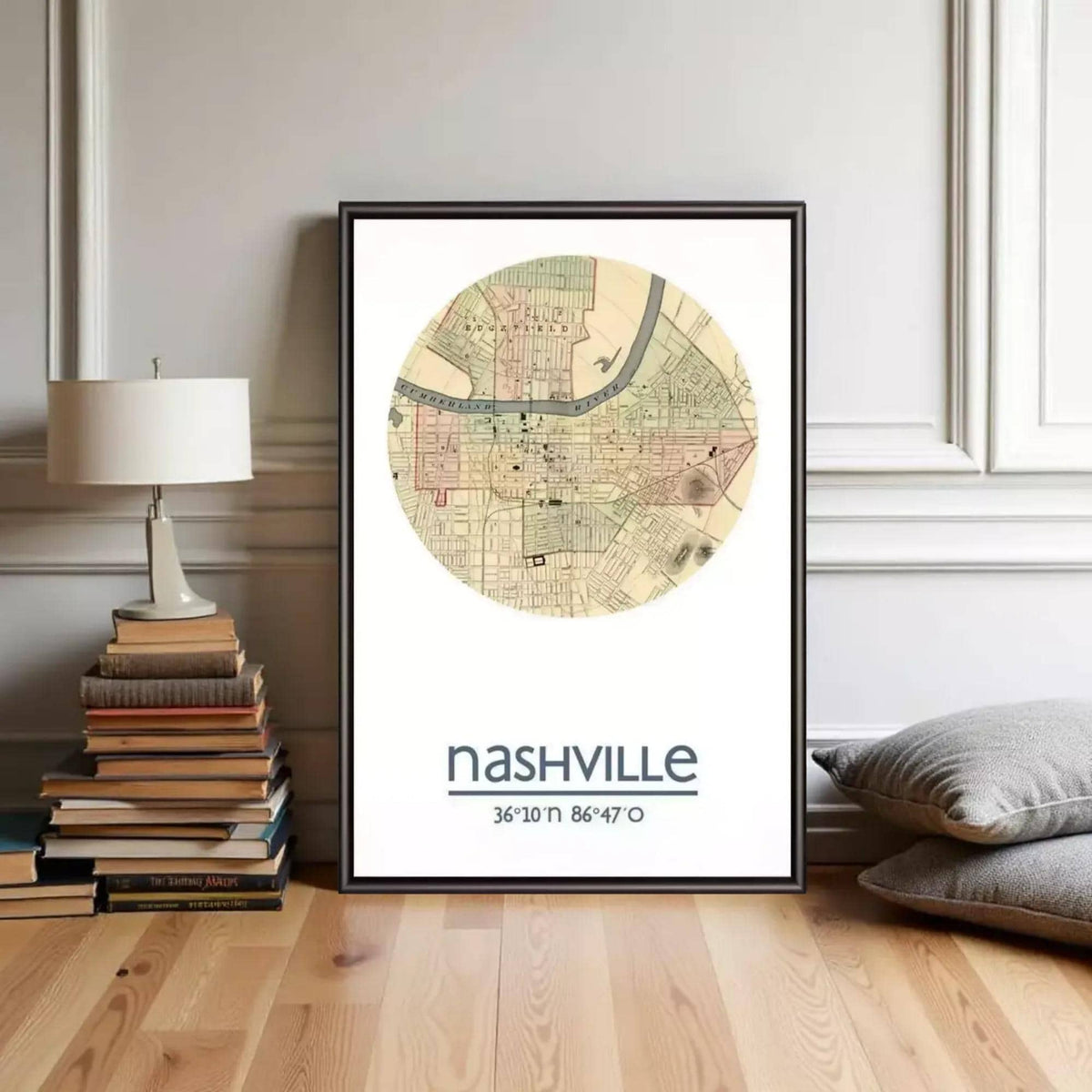 A framed map of Nashville, Tennessee, with the text " Nashville 36°10'11" 86°47'7"", placed on a wooden floor next to a stack of books and a lamp.