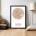 A framed map of Gdansk, Poland, with the city's name and location displayed prominently. The map is surrounded by a circular design, and the text "Gdansk" is written below it. The image is set against a white wall, with a lamp and some books visible in the background.