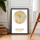 A framed map of Australia with the text "adelaide" printed on it, placed on a wooden floor next to a stack of books and a lamp.