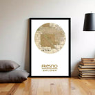 A framed map of Fresno, California, with the text "FRESNO" and "36.44.47.119.46.7 W" printed on it, standing on a wooden floor next to a stack of books and a lamp.