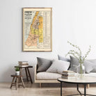 a living room with a framed vintage map of Palestine hanging on the wall above a couch with pillows. There are also some books and a vase with flowers on the couch, creating a cozy and inviting atmosphere.