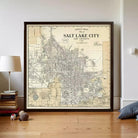a framed vintage map of Salt Lake City, Utah, with a lamp and some books on a wooden floor in the foreground.