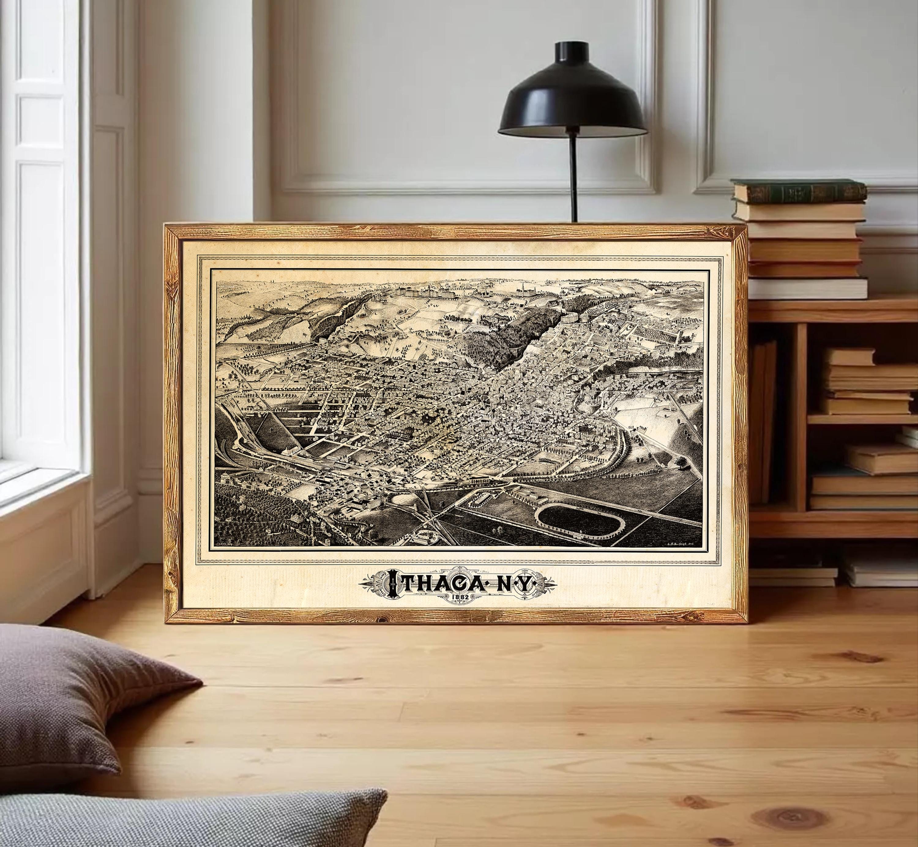 A framed black and white map of Ithaca, New York, displayed on a wooden floor in a room with a bookshelf and a lamp.