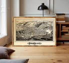 A framed black and white map of Ithaca, New York, displayed on a wooden floor in a room with a bookshelf and a lamp.