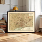 A framed map of Barcelona, Spain, displayed on a wooden floor in a room with a lamp and bookshelf in the background.