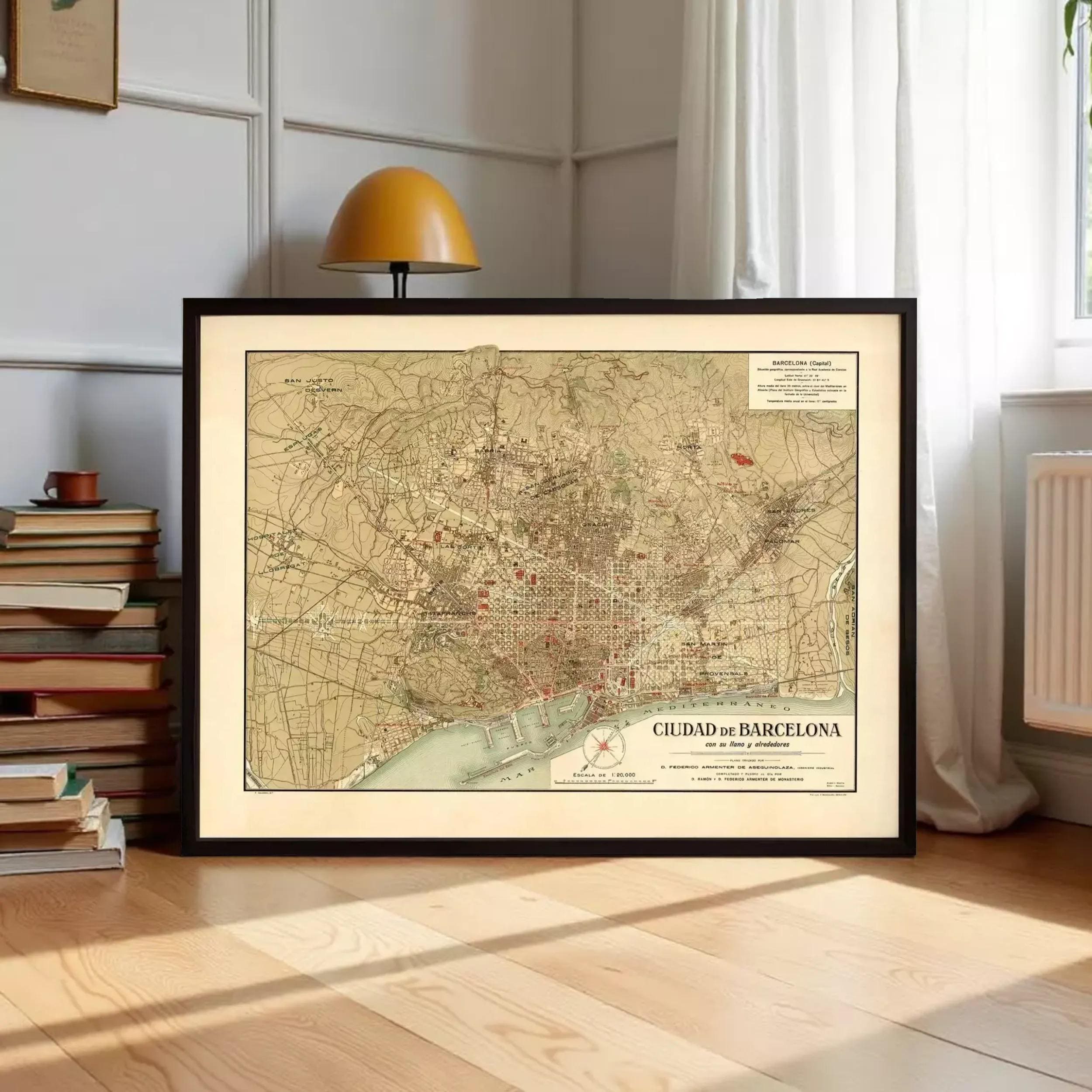 A framed map of Barcelona, Spain, displayed on a wooden floor in a room with a lamp and bookshelf in the background.