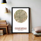 A framed map of Indianapolis, Indiana, with the city's name and location printed on it, is displayed on a wall in a room with a wooden floor and a stack of books.