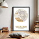 A framed map of Melbourne, Australia, with the text "melbourne 37° 47' 14'58" e" below it, is displayed on a wall in a room with a bookshelf and a lamp.