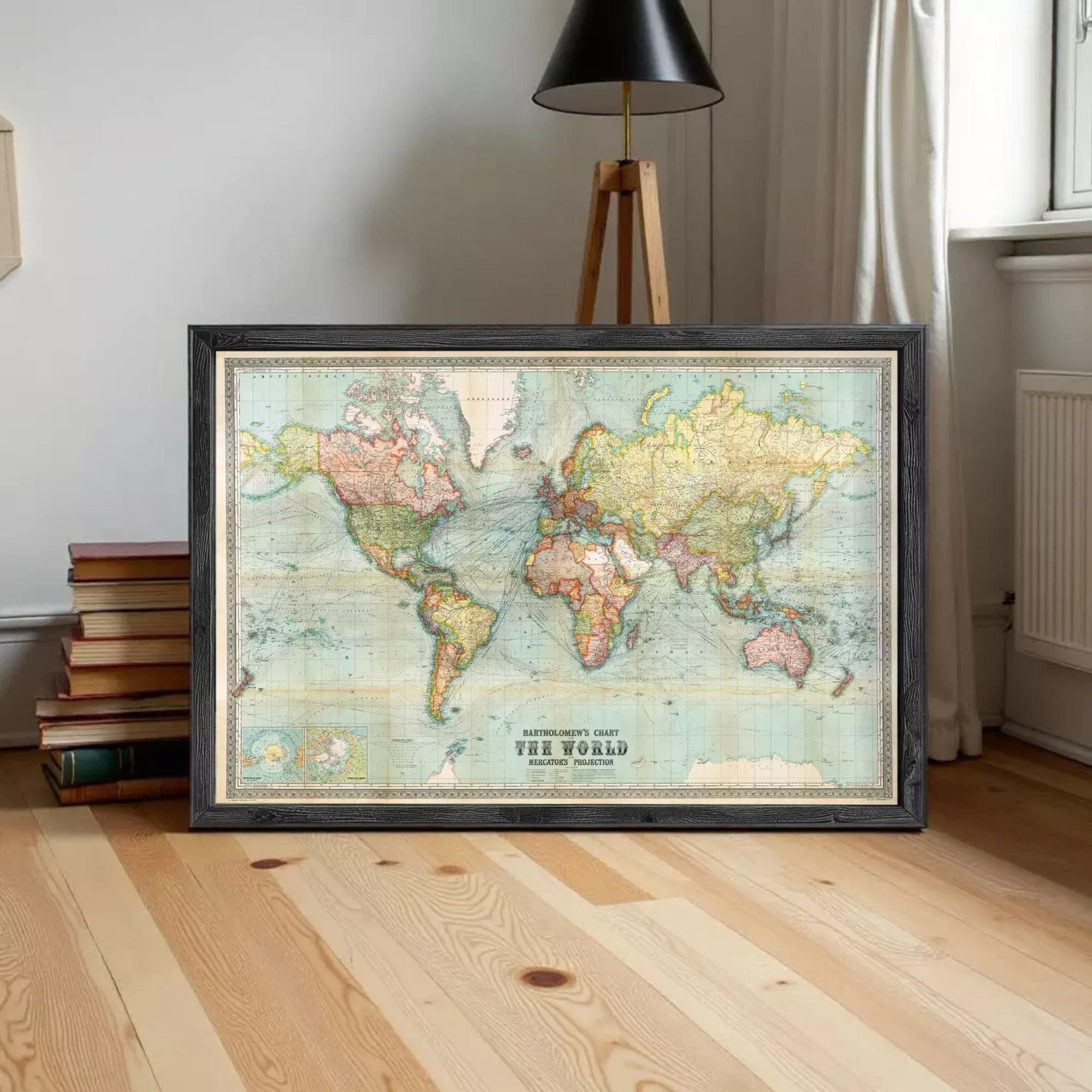 A framed vintage world map is displayed on a wooden floor in a room with a lamp and bookshelf in the background.