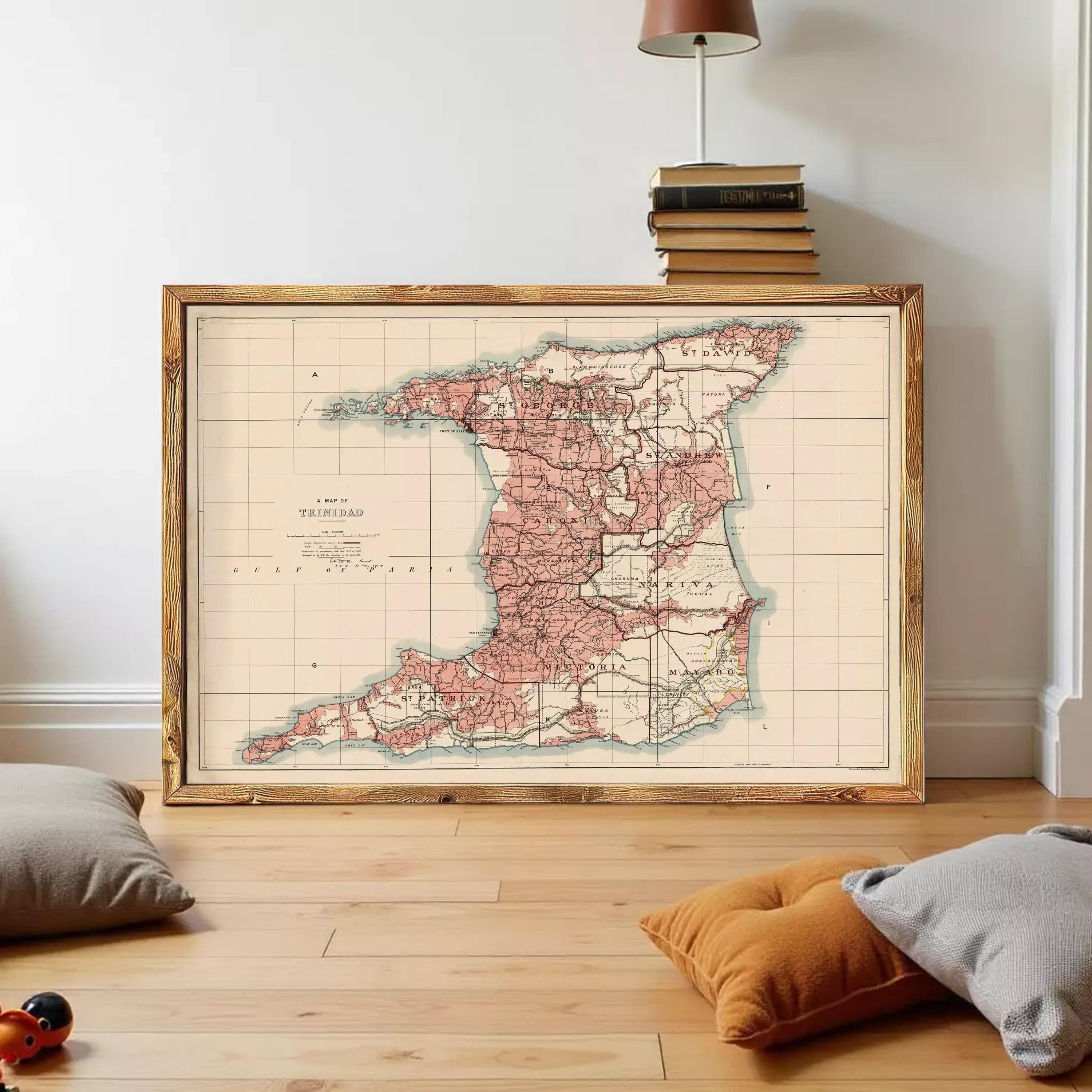a large framed map of Trinidad and Tobago, with a wooden frame and a vintage map design. The map is displayed on a wooden floor, with a stack of books and a lamp visible in the background.