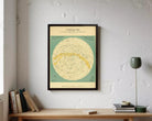a framed vintage-style map of the night sky, hanging on a wall above a wooden desk with books and a lamp.