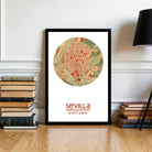 A framed map of Seville, Spain, with the text "seville" and "37.22°N 5.99°W" printed on it, is displayed on a wall next to a stack of books.
