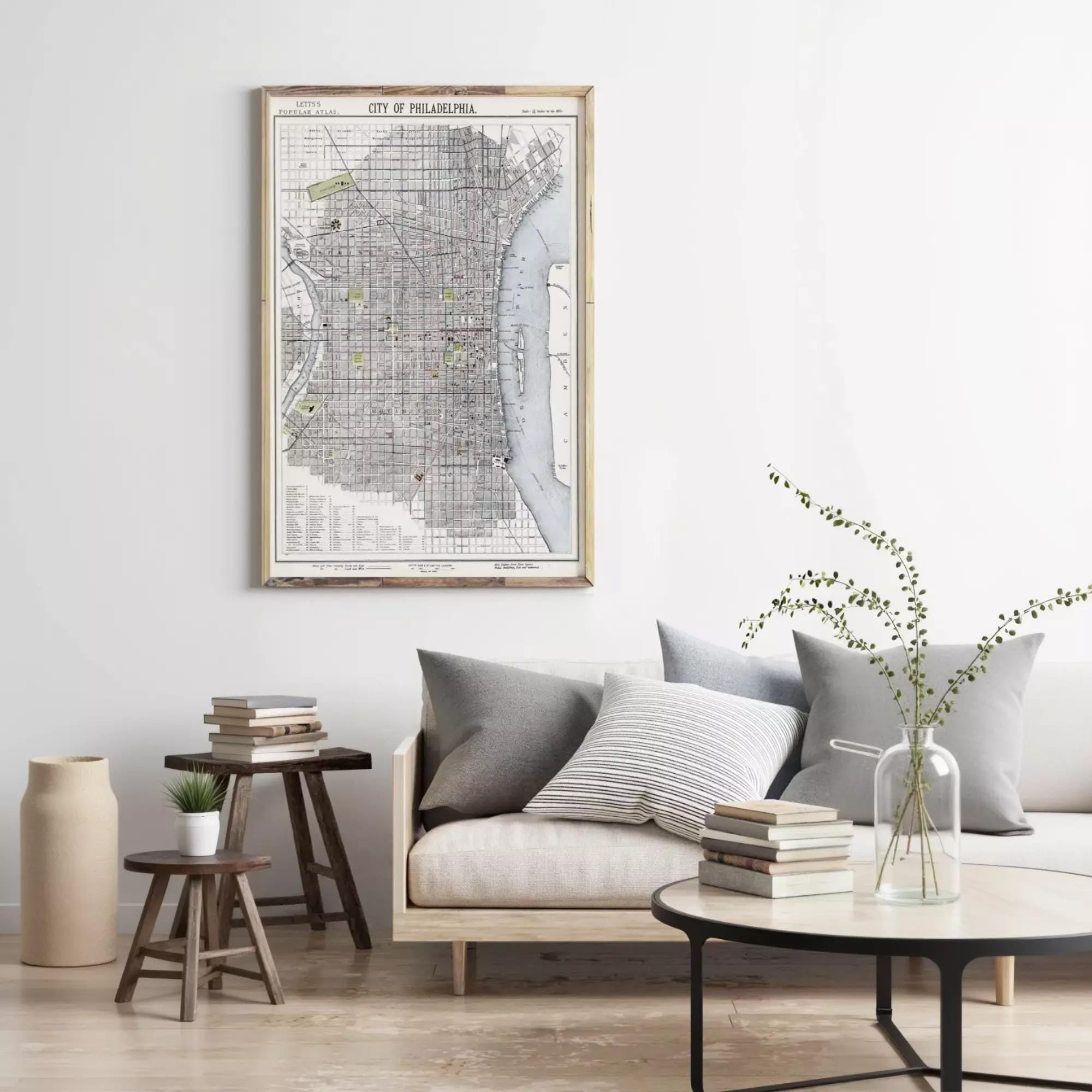 a living room with a large framed map of Philadelphia hanging on the wall above a couch with pillows. There are also some books and a vase with flowers on the couch, creating a cozy and inviting atmosphere.