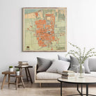 a living room with a large framed map of a city hanging on the wall above a couch with pillows. There are also some books and a vase with greenery on the coffee table in front of the couch.