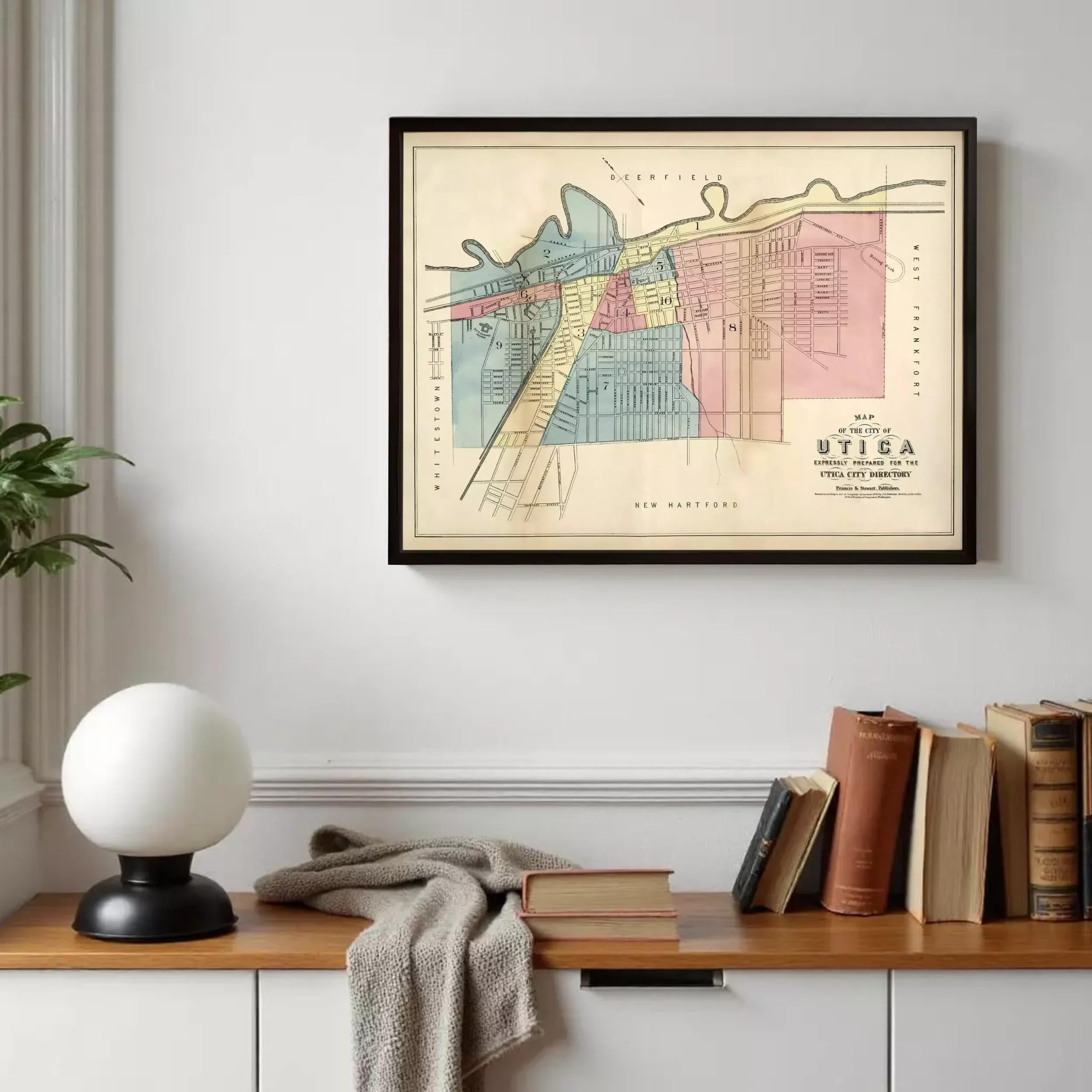a framed vintage map of the city of Utica, New York, hanging on a wall above a wooden bookshelf filled with books.
