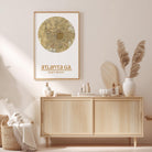a framed map of Atlanta, Georgia, hanging on a wall above a wooden cabinet. The cabinet has various items on top of it, including a vase with pampas grass, a bottle, and a book.