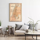 a living room with a framed map of Palestine hanging on the wall above a couch with pillows. There are also some books and a vase with plants on the couch, creating a cozy and inviting atmosphere.