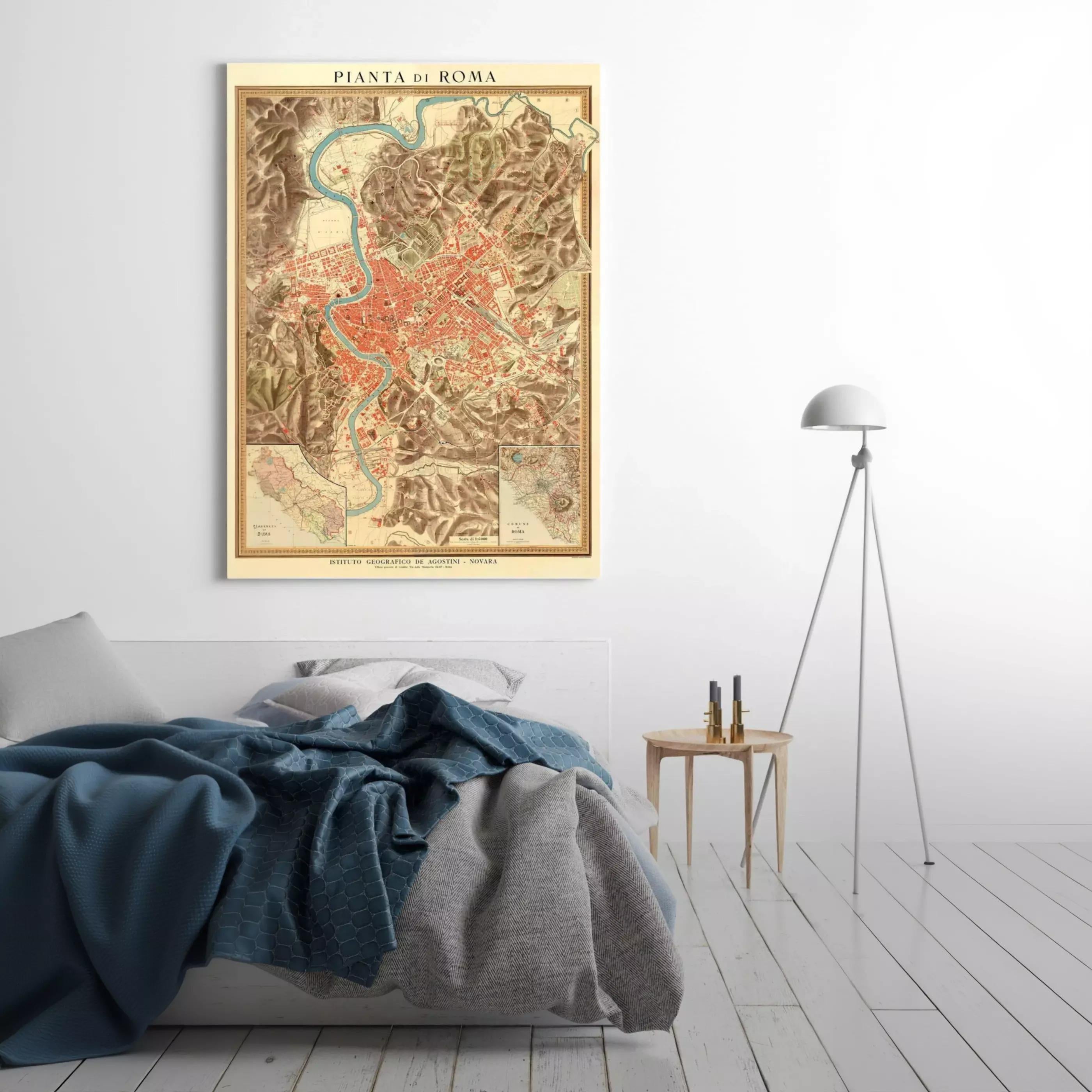 a bedroom setting with a bed, a nightstand, and a lamp. The bed has a blue blanket and a gray blanket on it. The nightstand has a lamp on it. The lamp is positioned next to the bed. The wall behind the bed has a framed map of Rome, which is titled "Pieta di Roma".