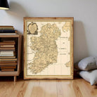 A framed vintage map of Ireland is displayed on a wooden floor next to a stack of books and pillows.