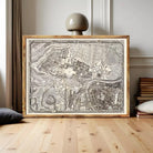 A large framed map of a city or region, with intricate details and a vintage appearance, displayed on a wooden floor in a room with books and a lamp.