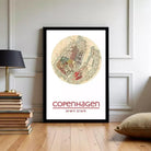 A framed poster of a map of Copenhagen, Denmark, with the text "COPENHAGEN" and "54°N 12°34°E" printed on it, displayed on a wall in a room with a stack of books and a lamp.