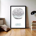 A framed poster of a map of Lisbon, Portugal, displayed on a wall in a room with a stack of books and a lamp.