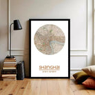 A framed map of Shanghai, China, with the text "Shanghai" and the date "31.11.21" and "21.28.20" printed on it, is displayed on a wall in a room with a lamp, books, and pillows.