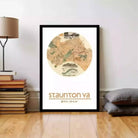 A framed map of Staunton, Virginia, with the text "Staunton VA 3897-79 W" printed on it, is displayed on a wall next to a stack of books and a lamp.