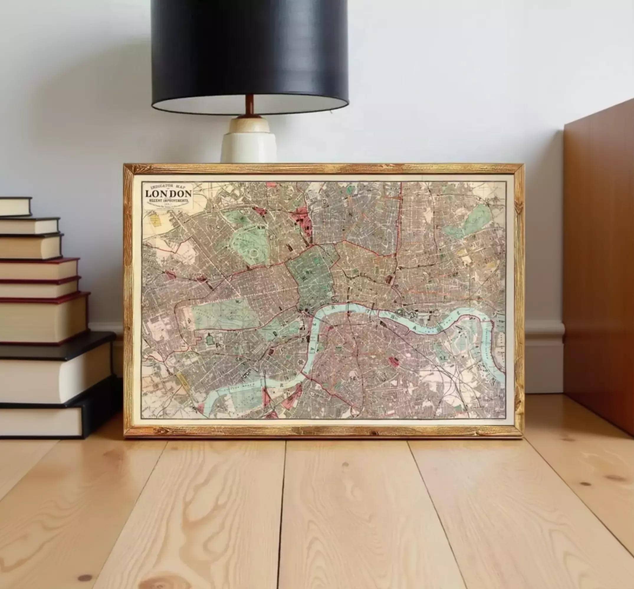 A framed vintage map of London is displayed on a wooden floor, with a lamp and books visible in the background.