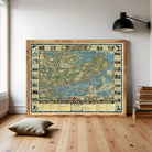a large framed map of the world, with various illustrations and labels, displayed on a wall in a room with a wooden floor and a stack of books nearby.