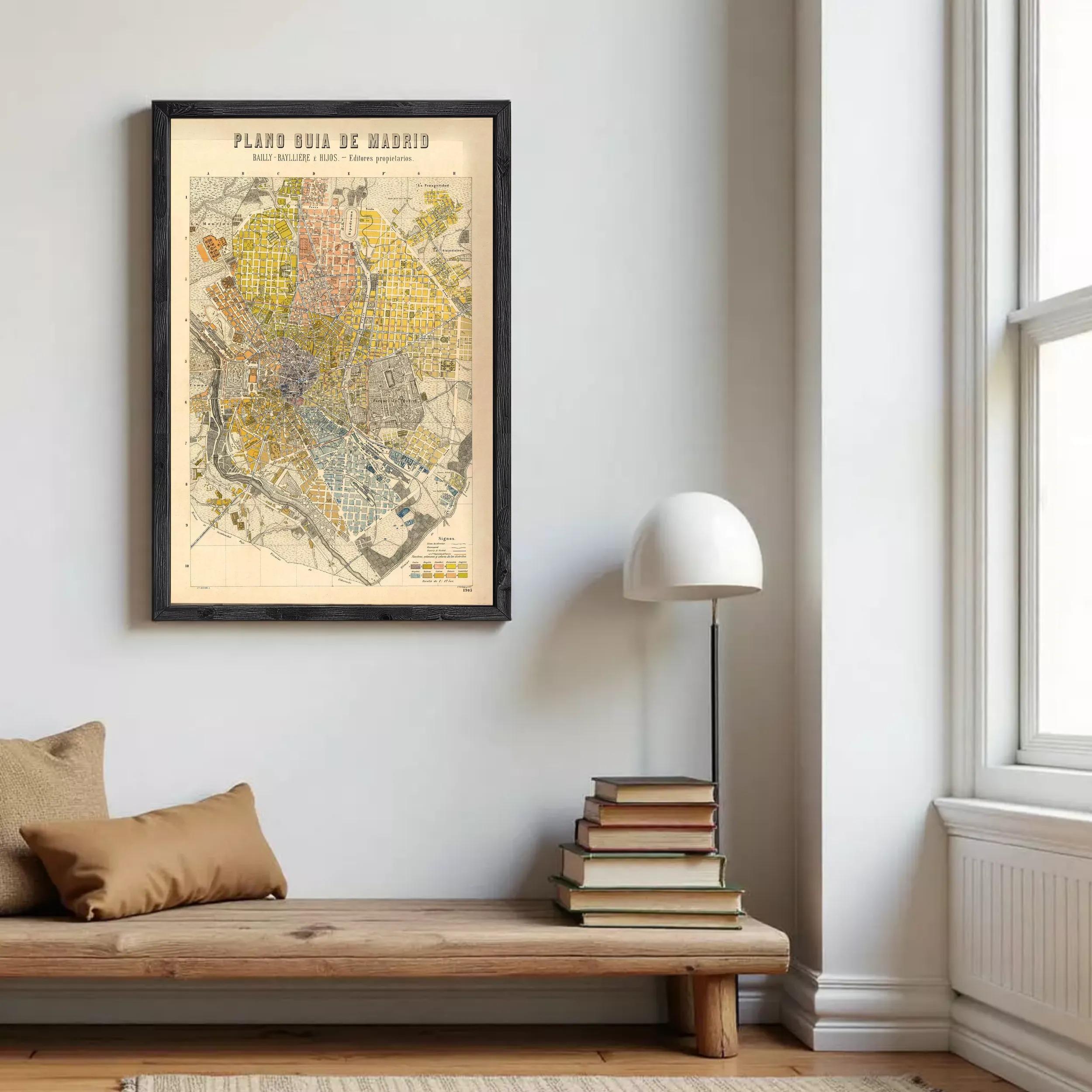 a framed map of Madrid hanging on a wall, with a stack of books and a lamp placed next to it.