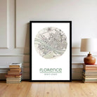A framed map of Florence, Italy, with the city's name and location displayed, is placed on a wall next to a stack of books and a lamp.