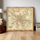 a large framed map of Hantaan, Germany, with a wooden frame and a vintage style. The map is displayed on a wooden floor, surrounded by a stack of books and a lamp.