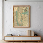 a framed vintage map of New York City hanging on a white wall above a wooden entertainment center with a stack of books and a teapot on top.
