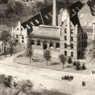 A black and white image depicting a large, multi-story brick building with a prominent chimney, surrounded by trees and a paved road.