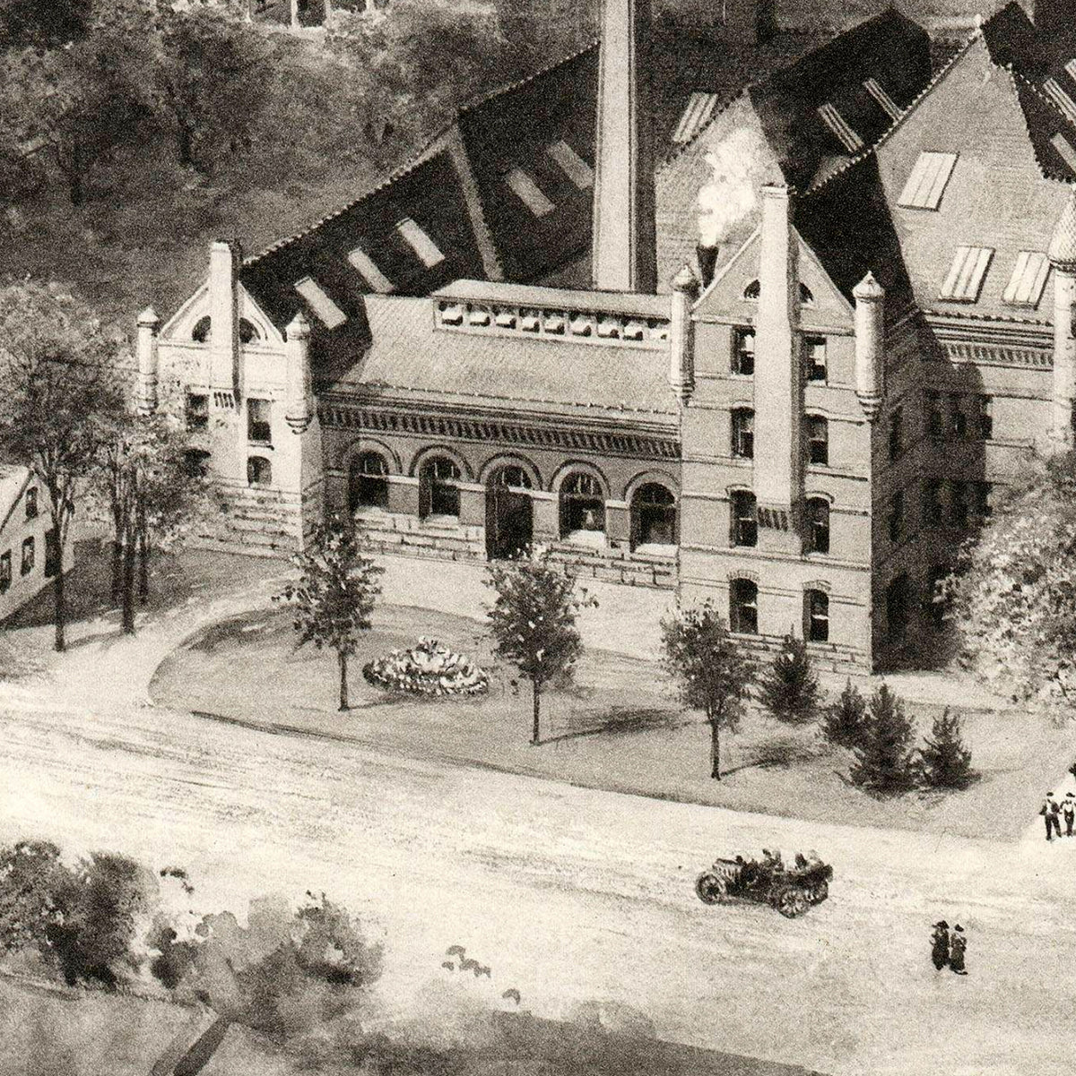 A black and white image depicting a large, multi-story brick building with a prominent chimney, surrounded by trees and a paved road.