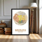 A framed poster of a colorful map of Barcelona, with the city's name and location prominently displayed. The poster is placed on a wooden floor next to a lamp and a stack of books.