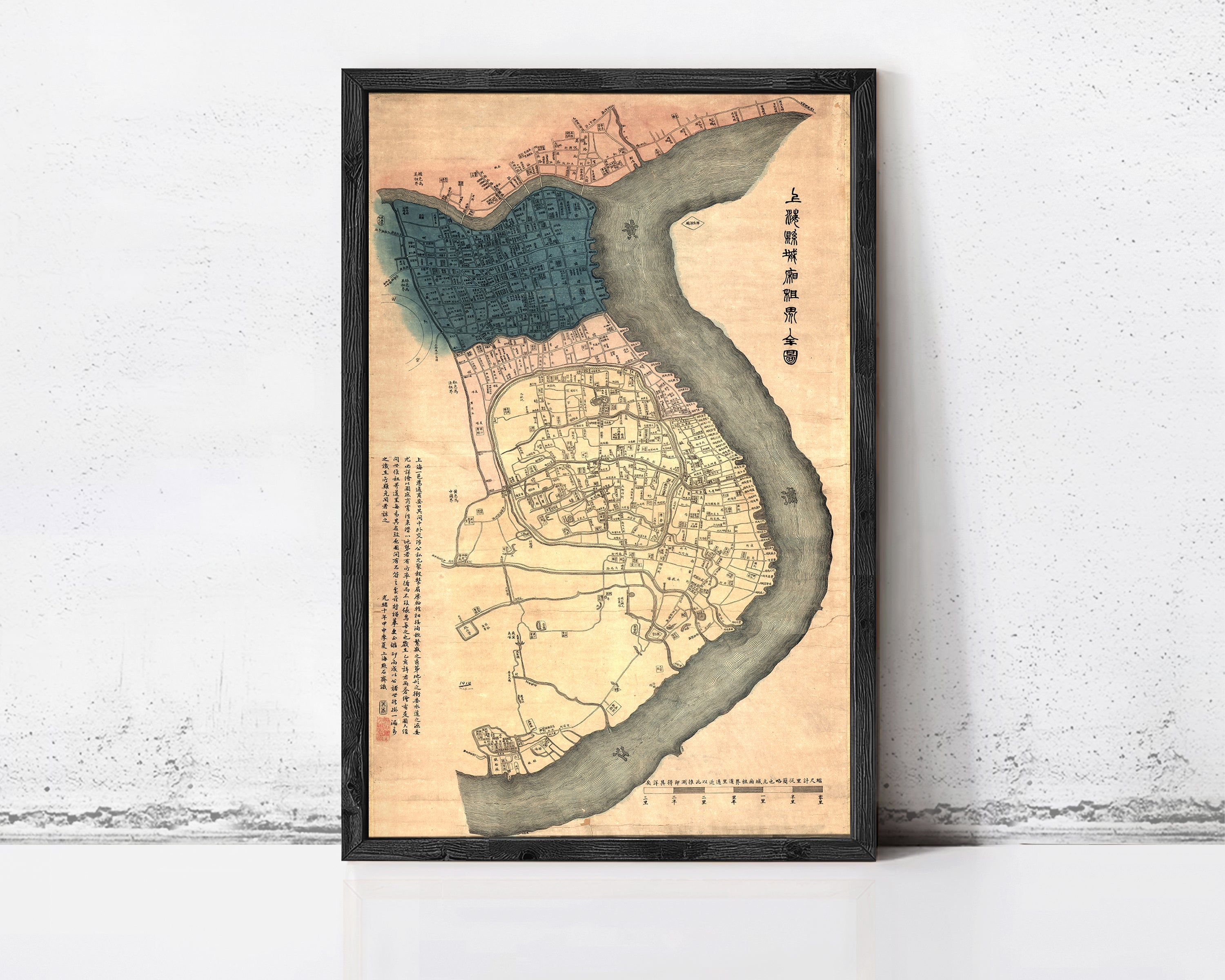The image is a framed map of a city, likely from an ancient Chinese or Japanese period, with a river running through it. The map appears to be in Chinese or Japanese script, and the frame has a black border. The map is displayed on a white wall, which contrasts with the colorful and detailed design of the map.