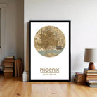 A framed map of Phoenix, Arizona, with the city's name and location printed on it, is displayed on a wall in a room with a stack of books and a lamp.