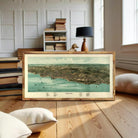 A framed map of Plymouth, Massachusetts, depicting the city's layout and surrounding areas, is displayed on a wooden floor in a room with bookshelves and a lamp.